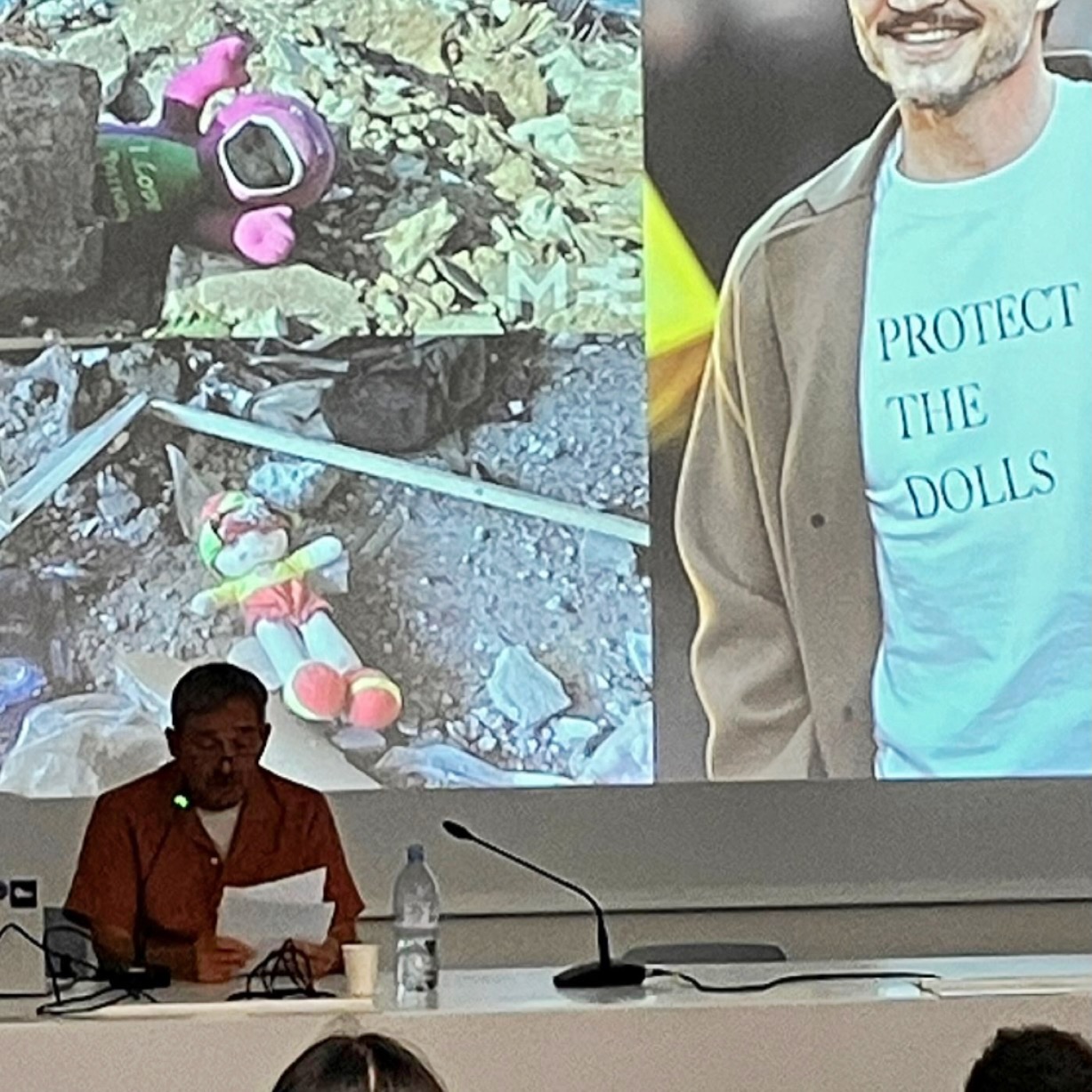 Photograph of me delivering my talk. I'm sitting at a desk and a behind me is projected two images of cuddly toys in the rubble of Gaza and one of Pedro Pascal wearing a t-shirt with the slogan 'Protect the Dolls.'