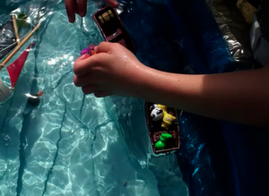 still from video essay, a child's hand and toys in a sunlit paddling pool