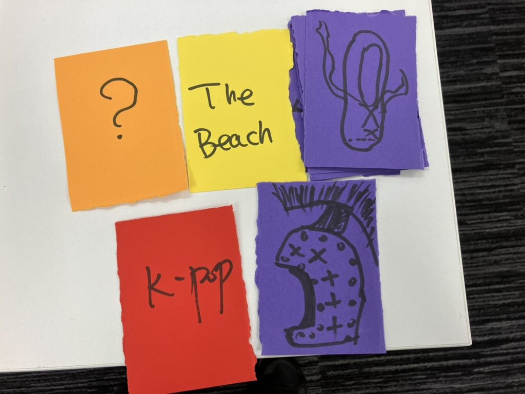 A prototype card game on brightly coloured paper. Cards carry the words and symbols 'K-Pop', '?', and drawings of a ballet shoe and a Roman helmet.