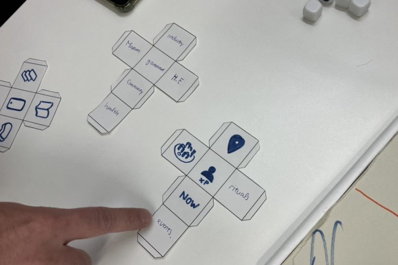 Finger pointing at a paper dice under construction with words and emblems on its surfaces