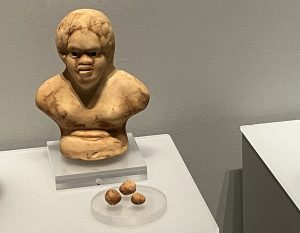 A child's rattle in the shape of a human torso and head. It is made from baked clay and has three of its beads next to it in its museum case.