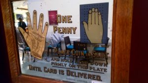 Part of an early twentieth century pier or arcade machine. The sign promises to read the user's palm for one penny, and gives instructions.