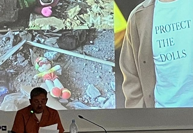 Photograph of me delivering my talk. I'm sitting at a desk and a behind me is projected two images of cuddly toys in the rubble of Gaza and one of Pedro Pascal wearing a t-shirt with the slogan 'Protect the Dolls.'