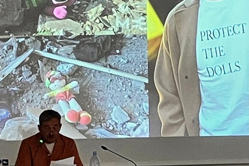 Photograph of me delivering my talk. I'm sitting at a desk and a behind me is projected two images of cuddly toys in the rubble of Gaza and one of Pedro Pascal wearing a t-shirt with the slogan 'Protect the Dolls.'