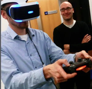 Photograph of a man in a VR headset with games controller watched by another man laughing