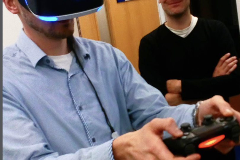 Photograph of a man in a VR headset with games controller watched by another man laughing